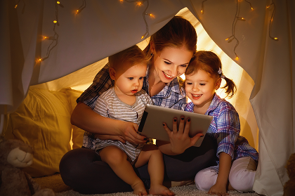 A mother and two children are sitting in a cozy fort, looking at a tablet together, surrounded by soft lighting and blankets.