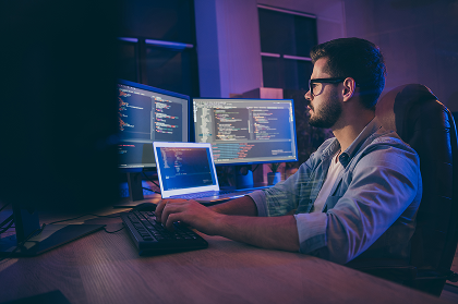 A man with glasses is coding at a desk with multiple computer screens displaying code in a dimly lit room.